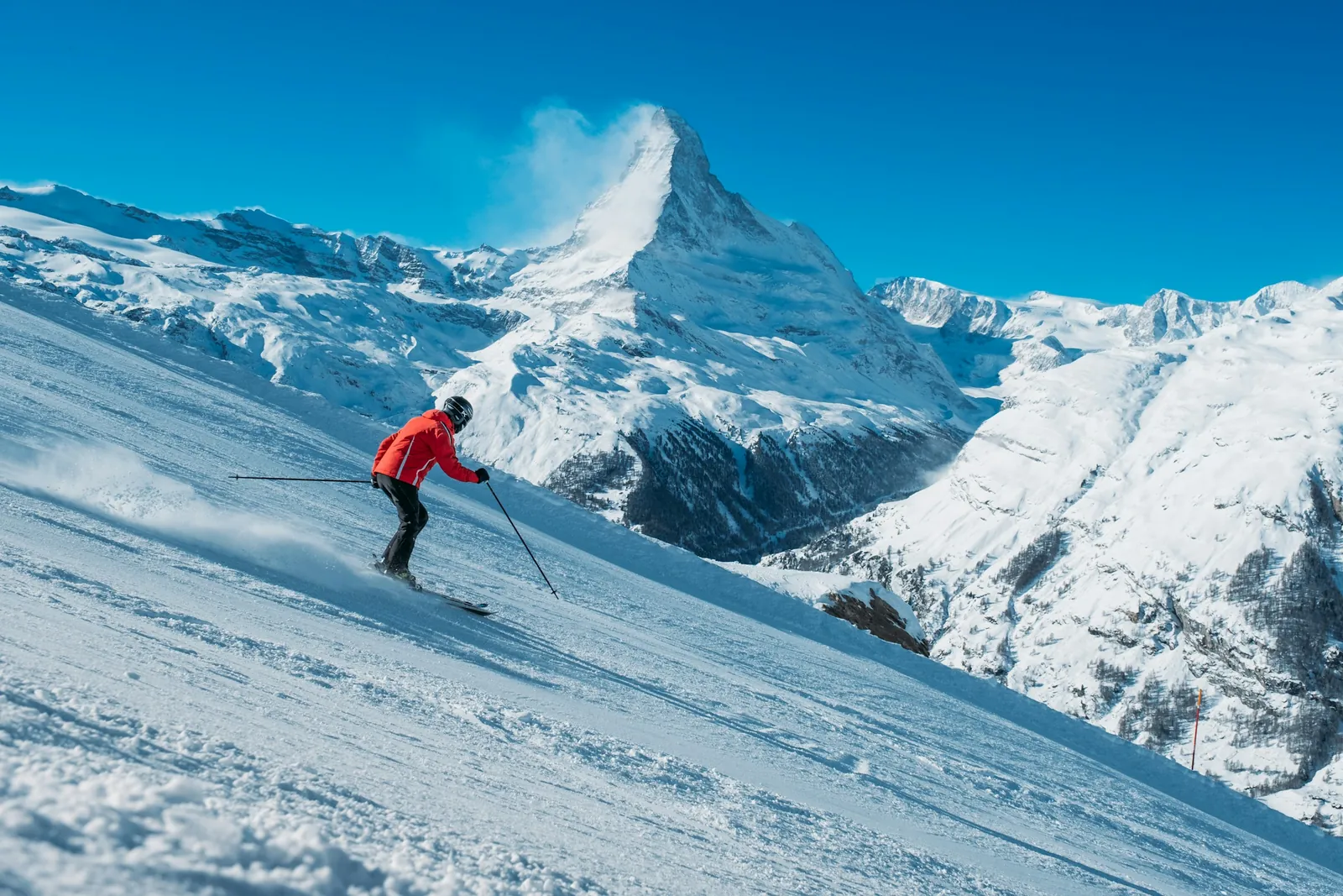 A skier in a red jacket carves down a sunny slope below the snow-covered Matterhorn.