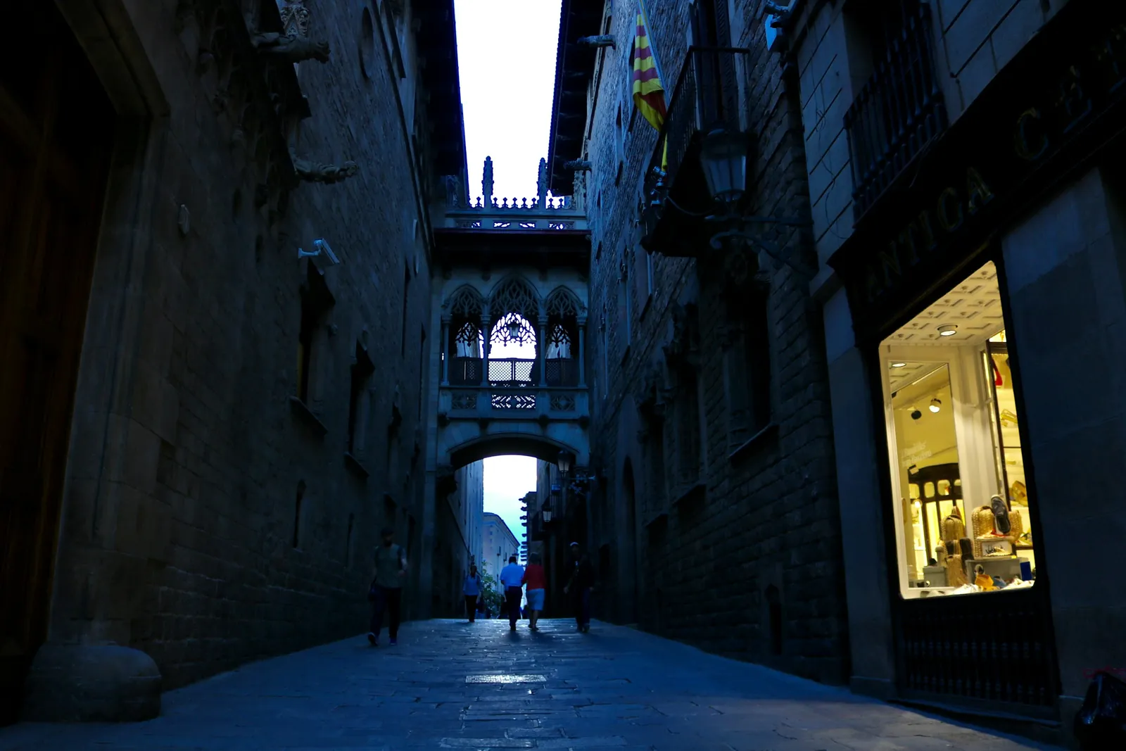 People walk down a narrow street in Barcelona's Gothic Quarter, under a stone bridge carved with Gothic arches.
