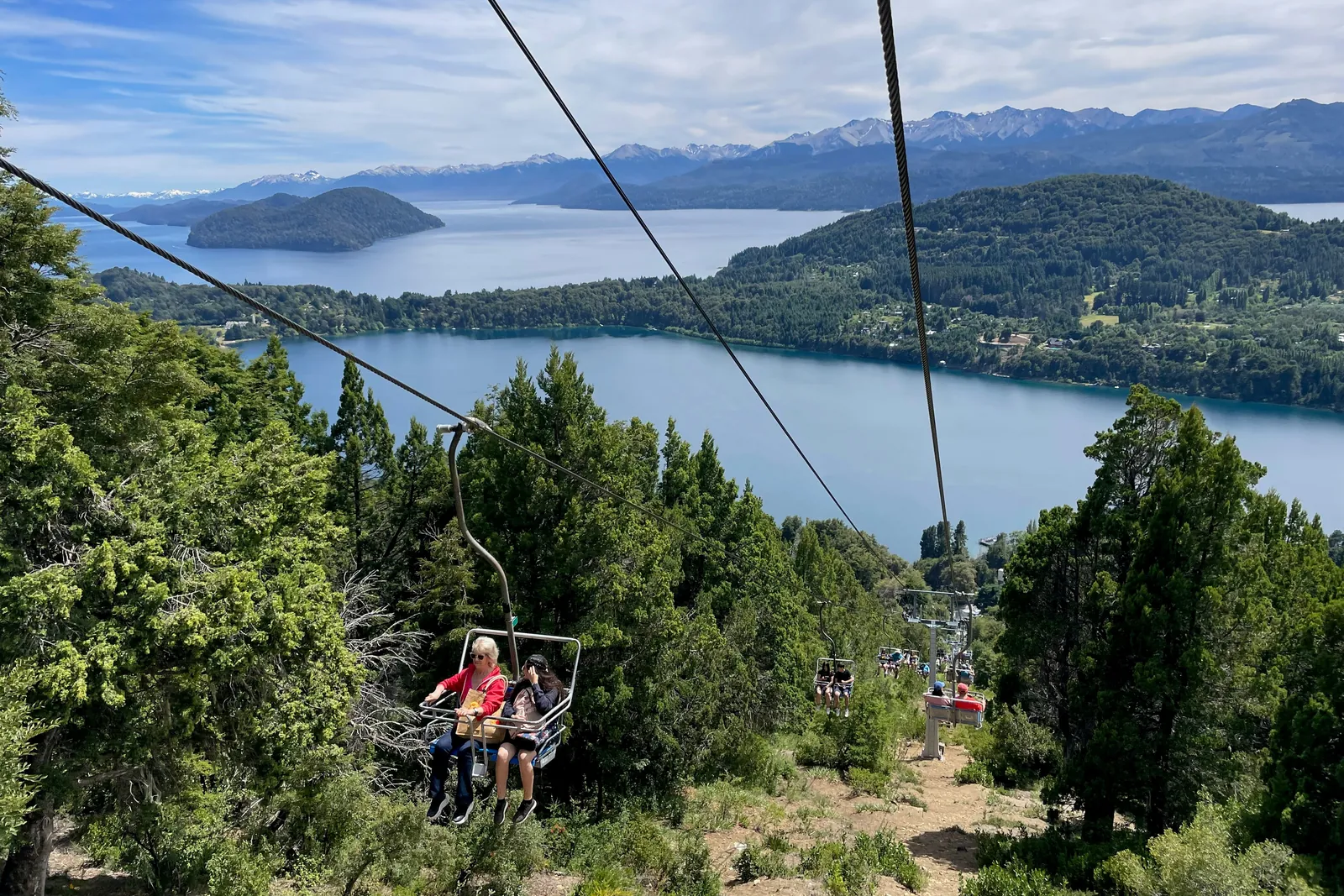 Two people ride a chairlift up a green, wooded hill above Bariloche's blue lakes, with snow-streaked mountains in the distance.