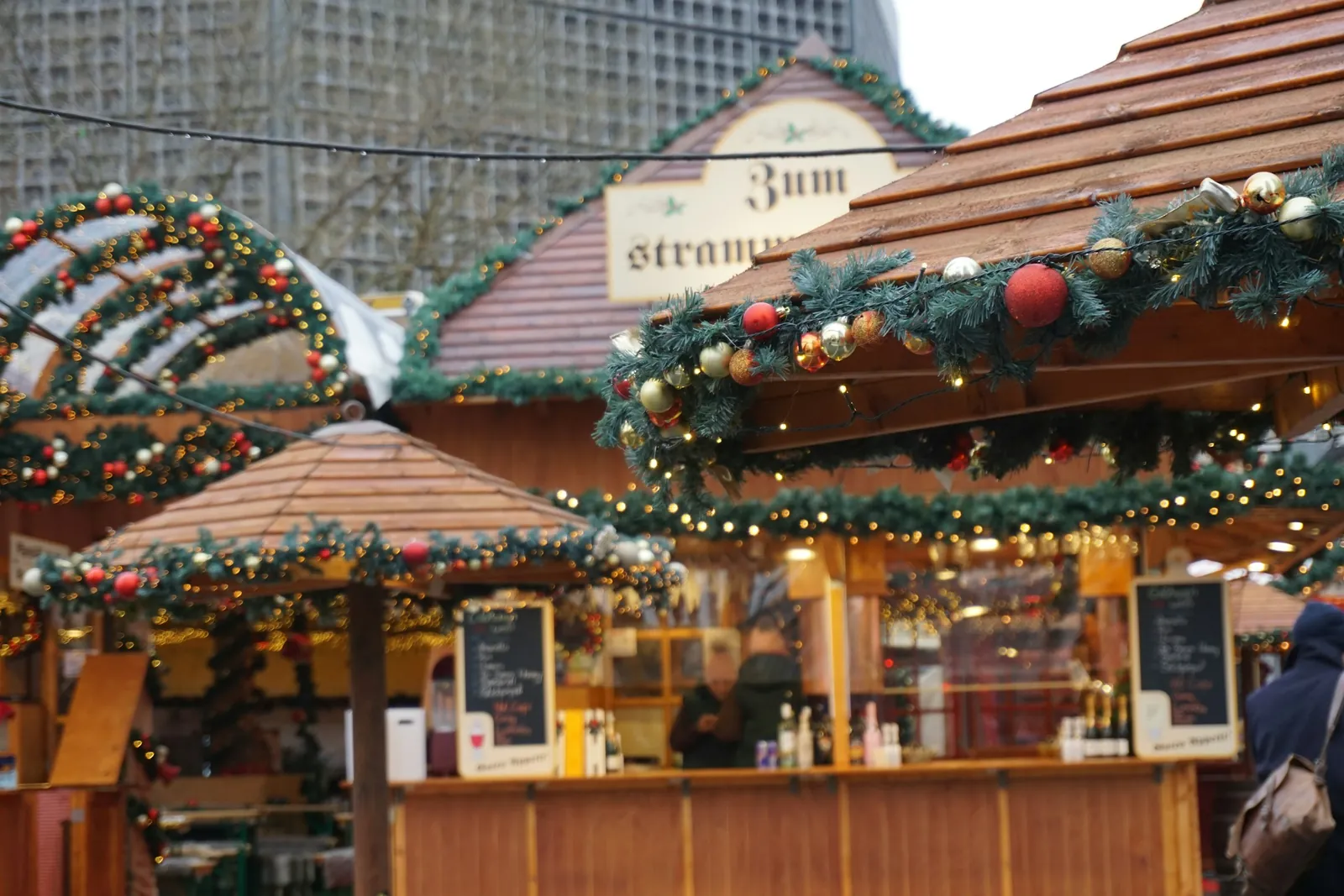 Uma barraca de madeira de um mercado de Natal em Berlim, enfeitada com guirlandas e bolas vermelhas, com pessoas no balcão.