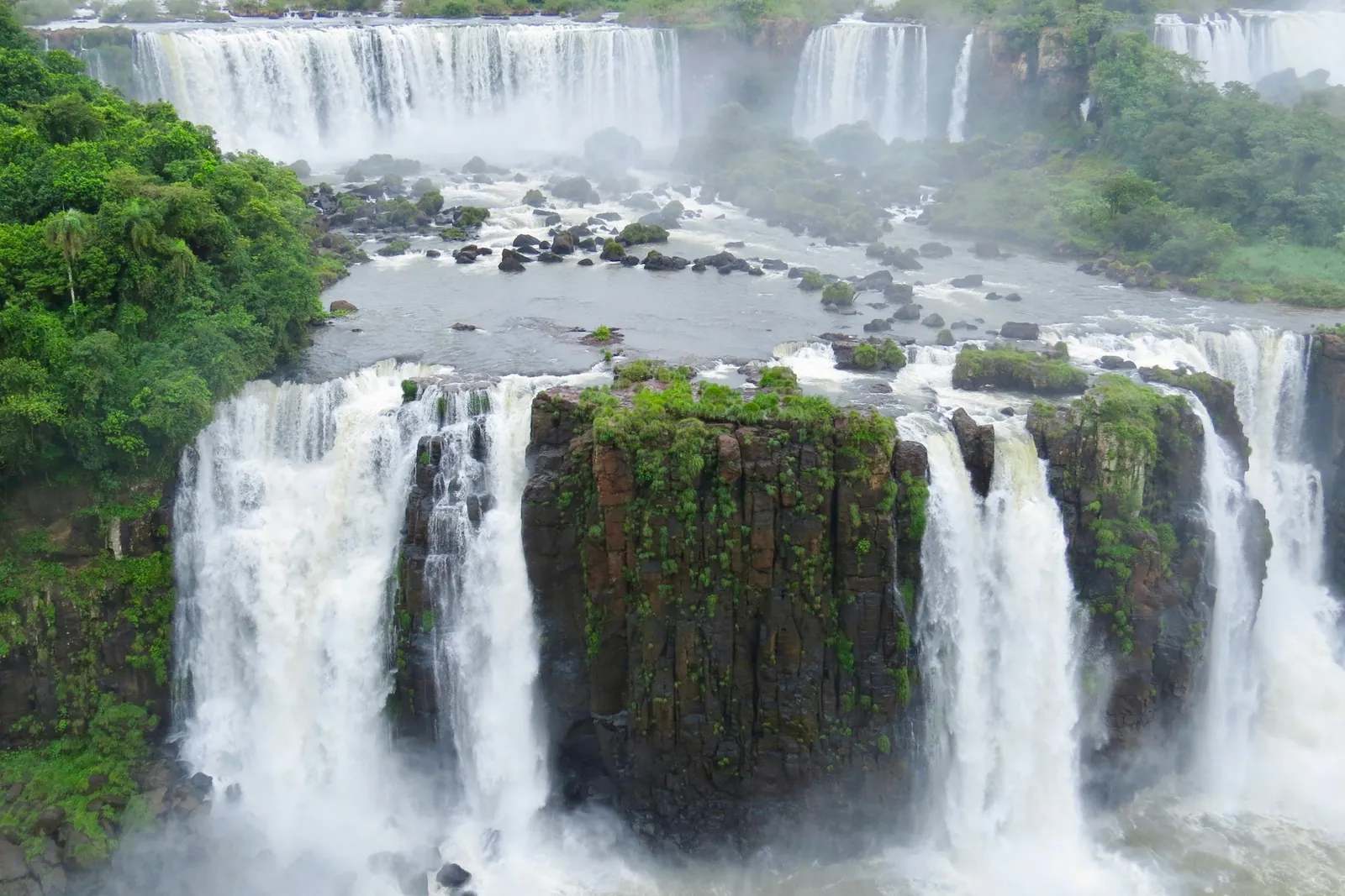 Varias caídas de agua se desploman desde paredones cubiertos de musgo en Iguazú, con más cascadas y selva al fondo.