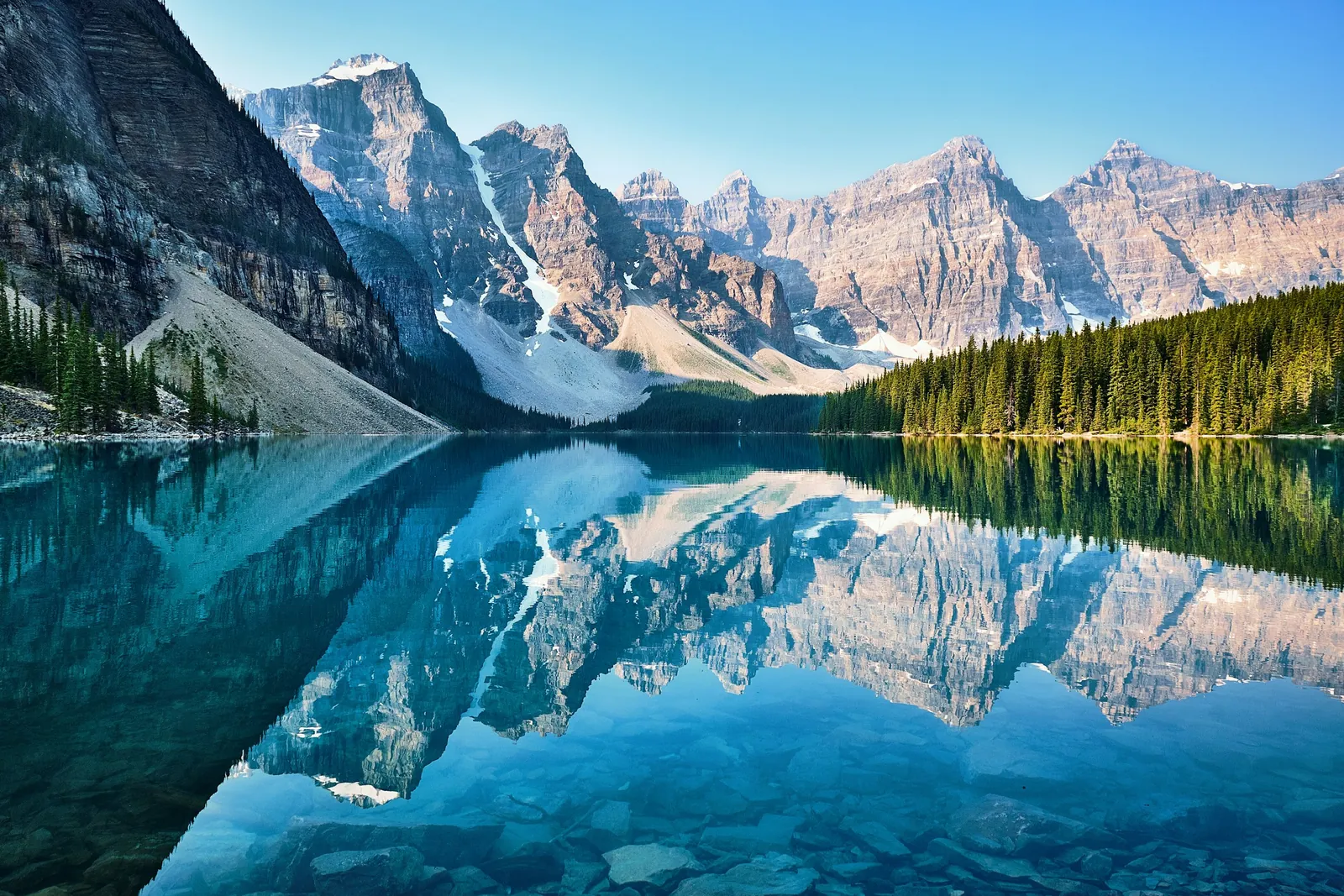The still, turquoise water of Moraine Lake mirrors the snowy peaks and pine forest around it.