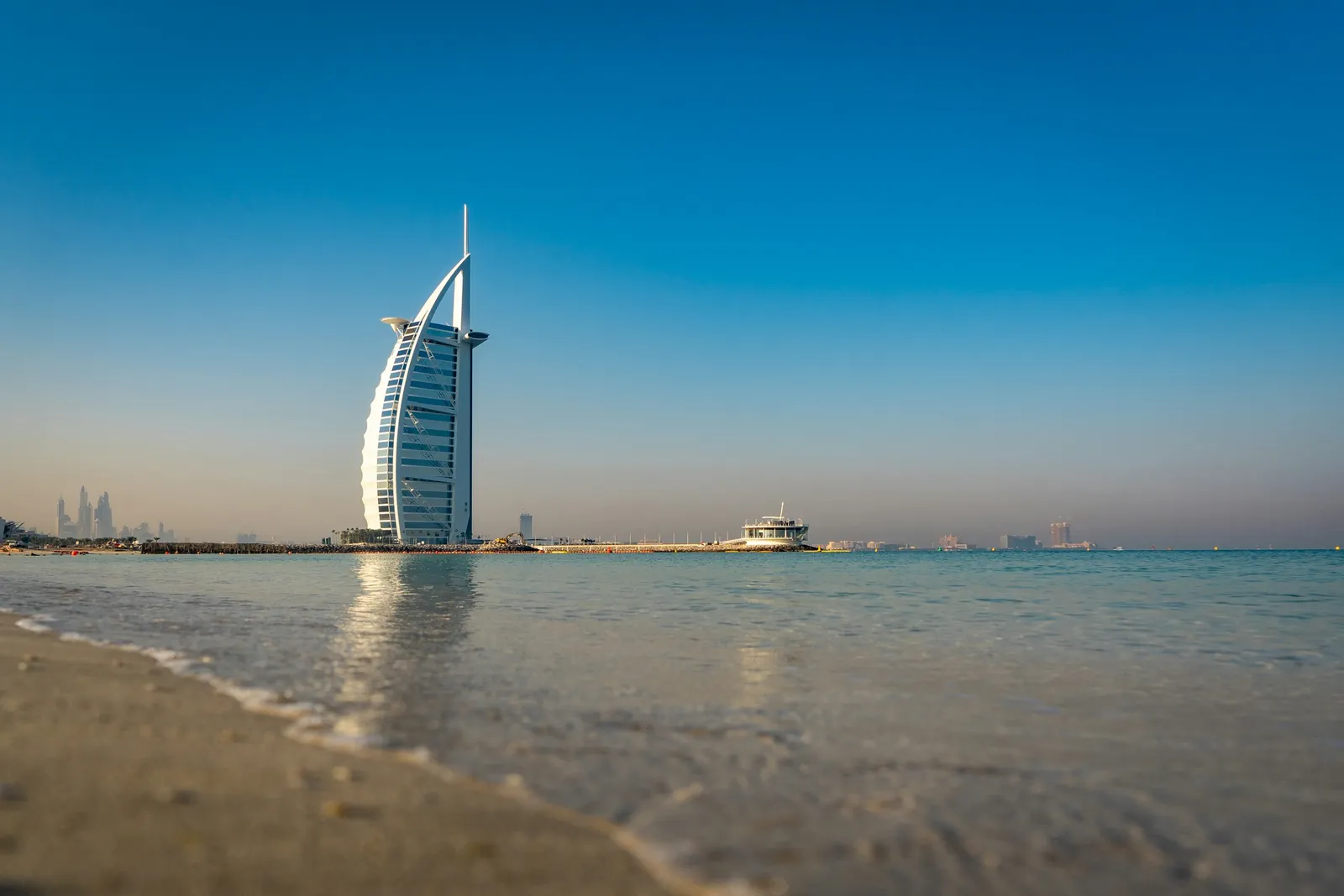 The sail-shaped Burj Al Arab rises from a calm sea, seen from the sand of a Dubai beach under a clear blue sky.