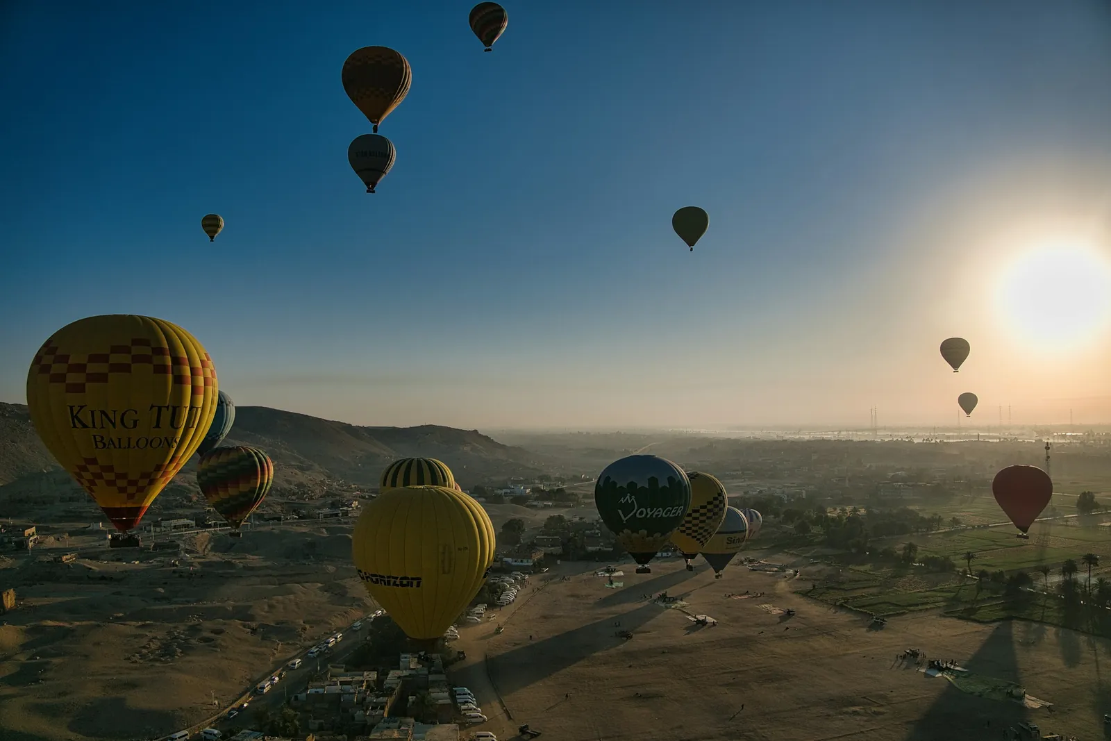 Globos aerostáticos se elevan al amanecer sobre las colinas del desierto y los campos verdes de la orilla oeste de Luxor, con el sol bajo en un cielo despejado.