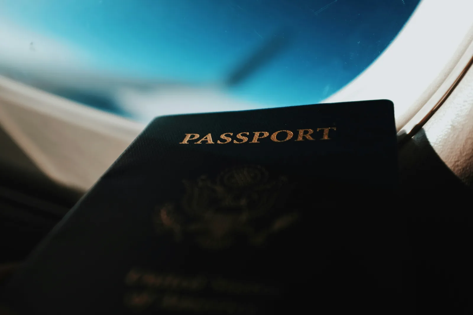 Un pasaporte oscuro sostenido frente a la ventanilla de un avión, con el cielo azul afuera.