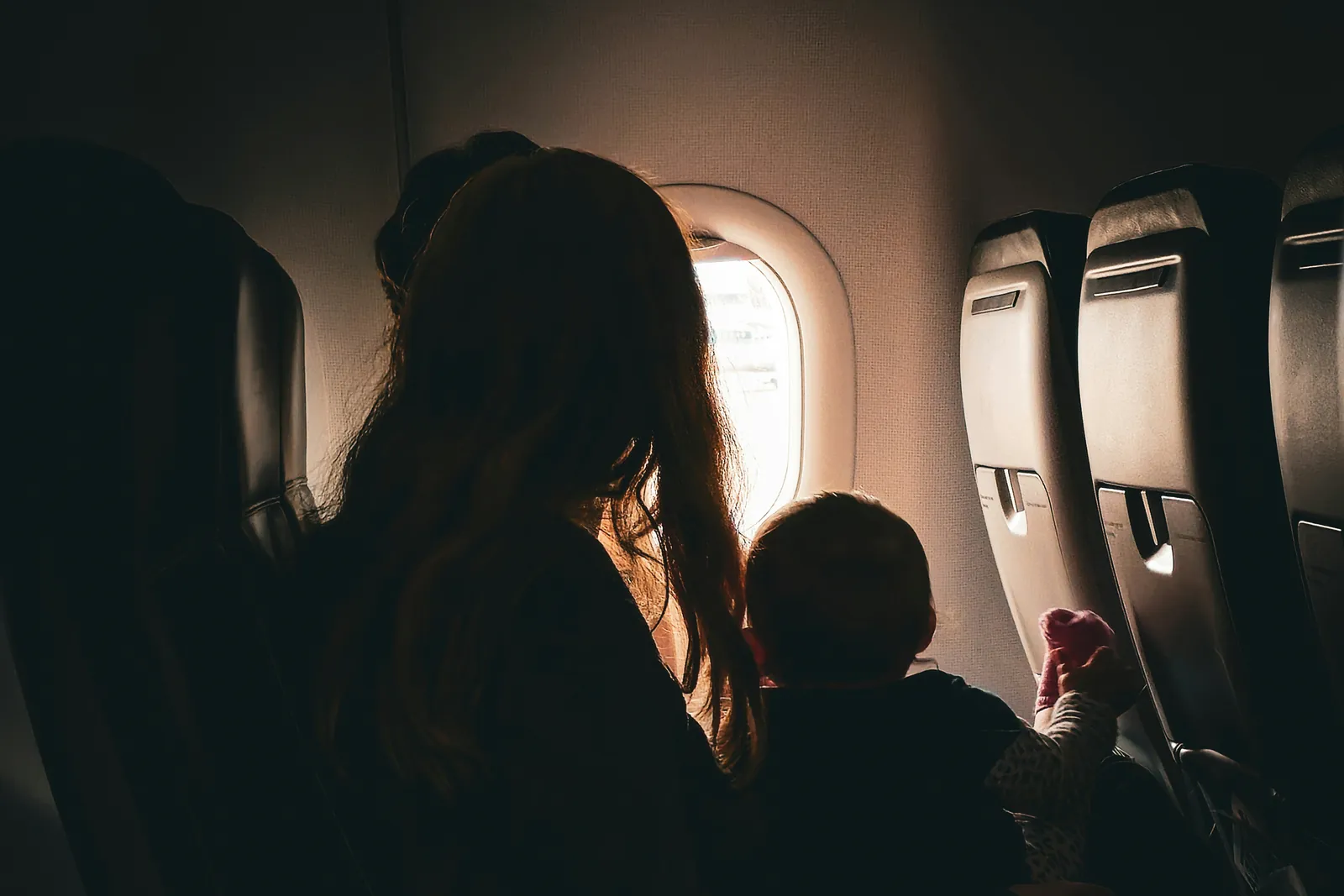 Na cabine escura de um avião, uma mãe e seu bebê, de costas, olham por uma janela iluminada.