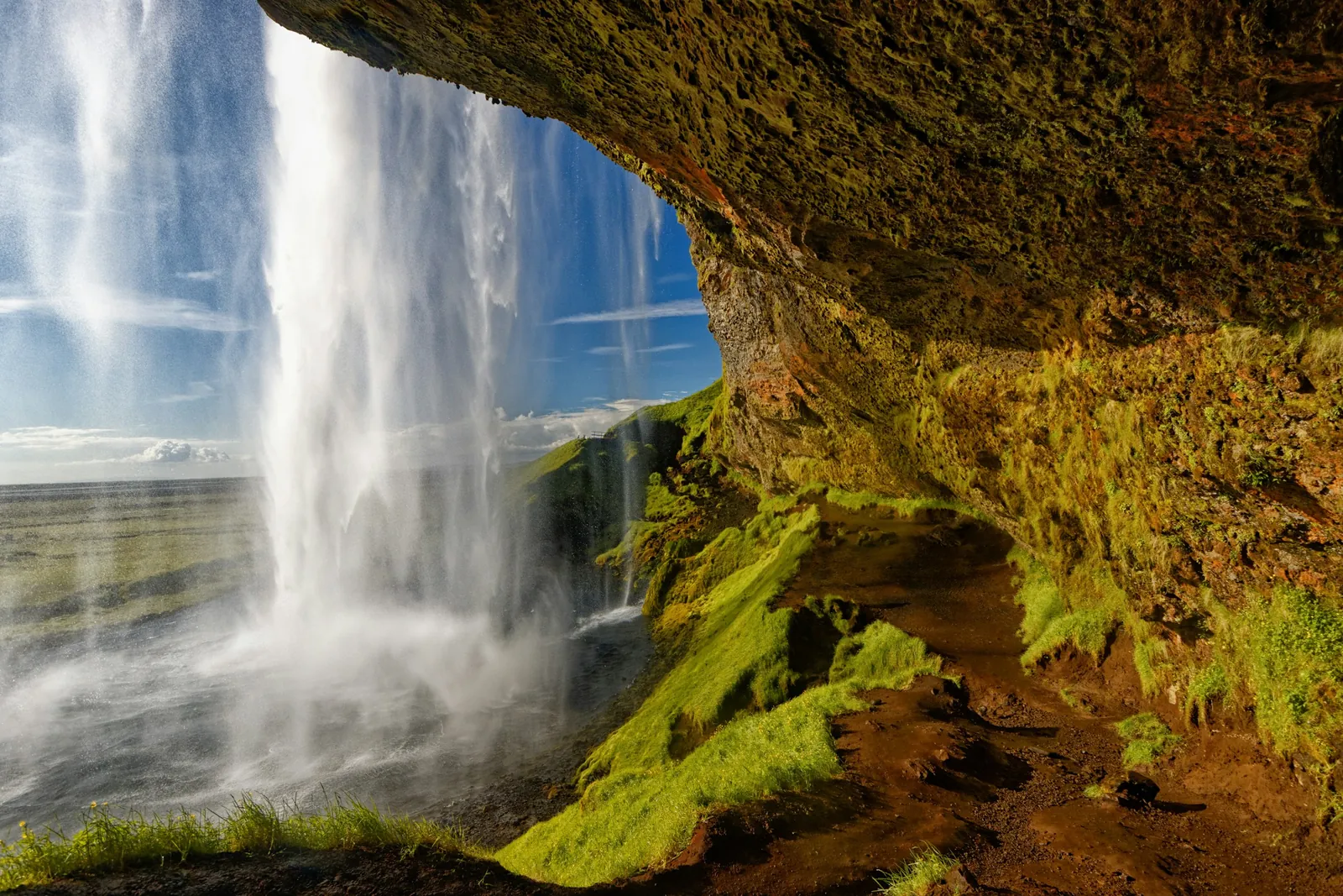 Seljalandsfoss falls past a mossy cave, seen from the path that runs behind the waterfall.