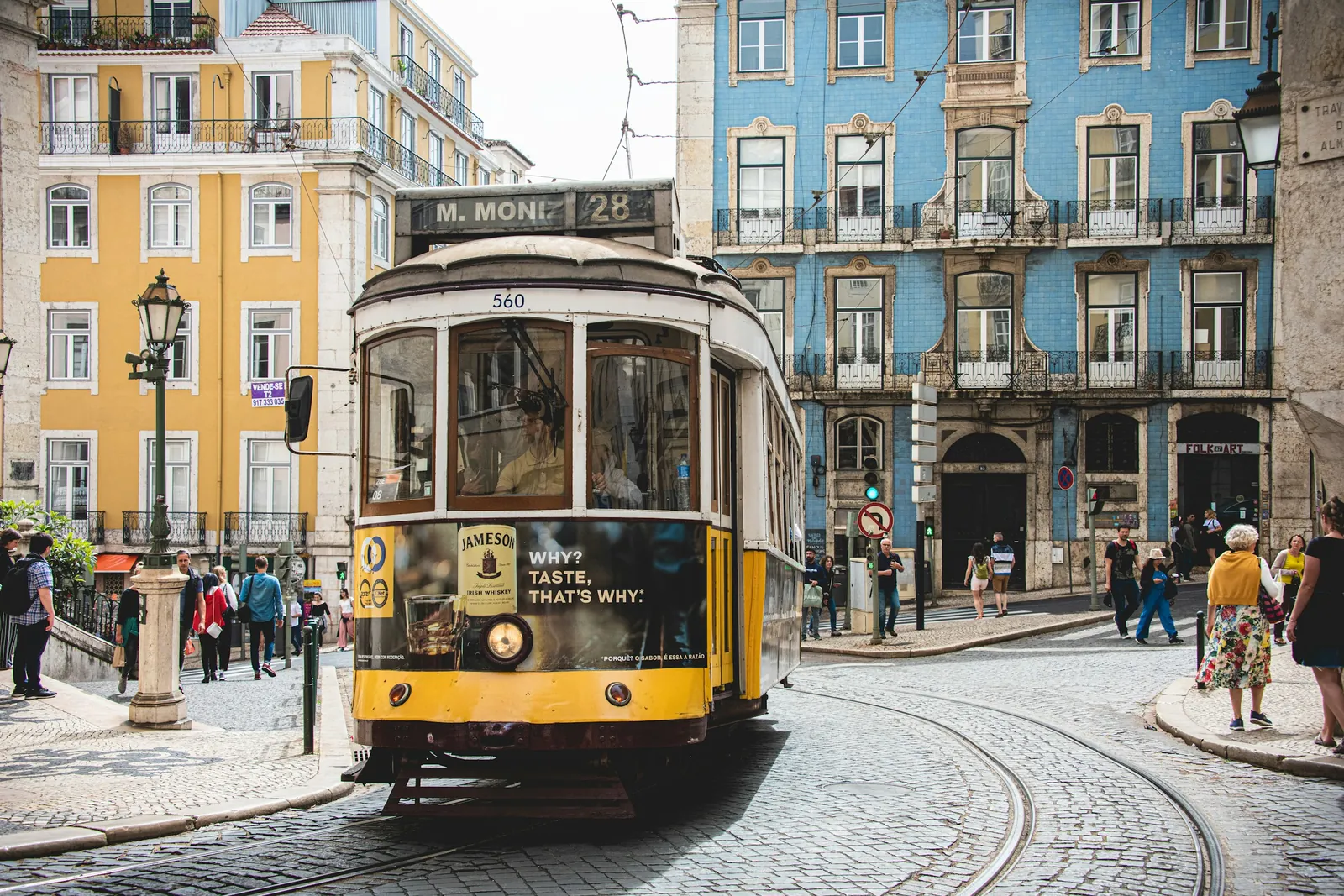 Um bonde amarelo da linha 28 atravessa uma praça de paralelepípedos em Lisboa, diante de prédios cobertos de azulejos azuis.