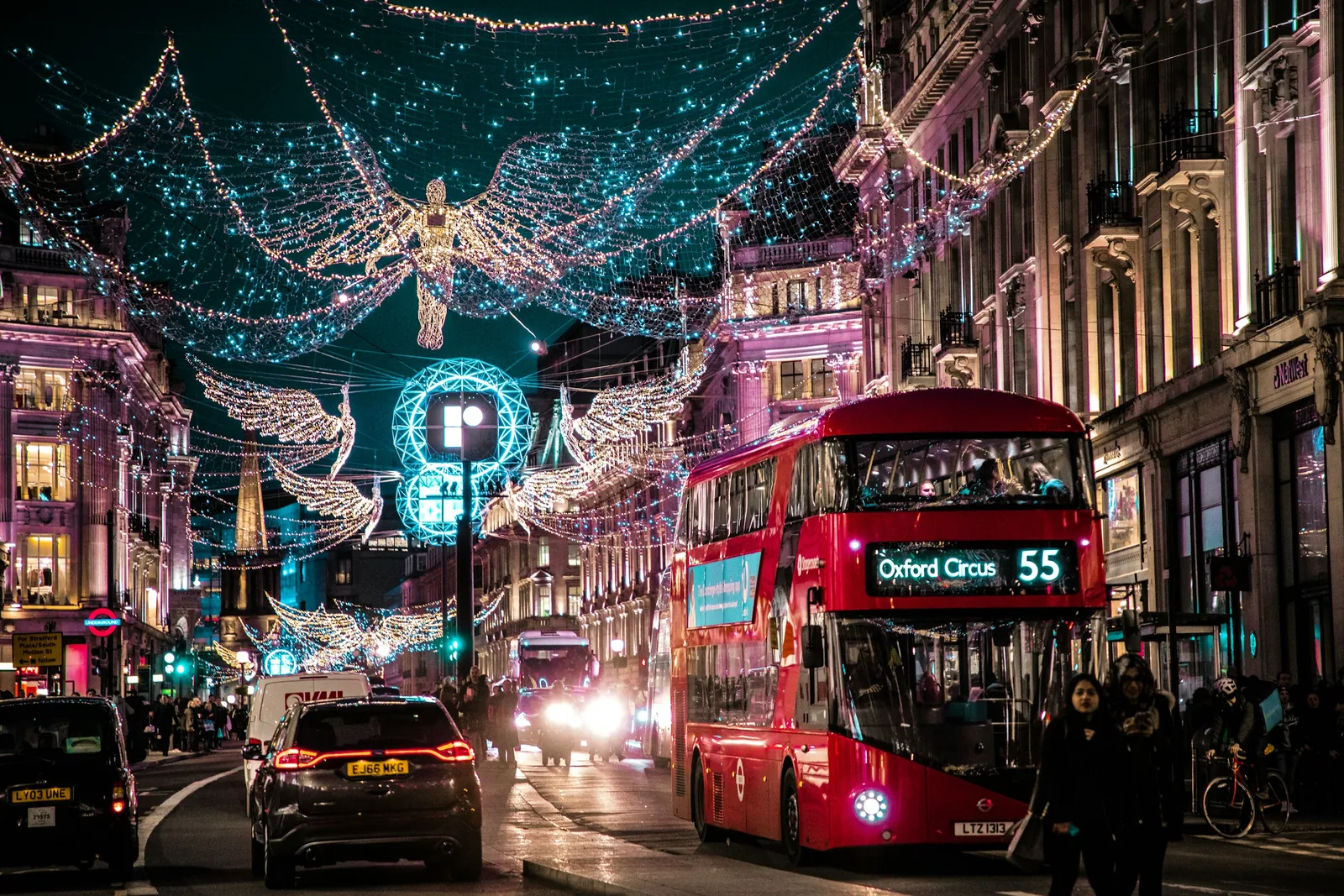 A red double-decker bus drives down Regent Street under Christmas lights shaped like angels, in the evening.