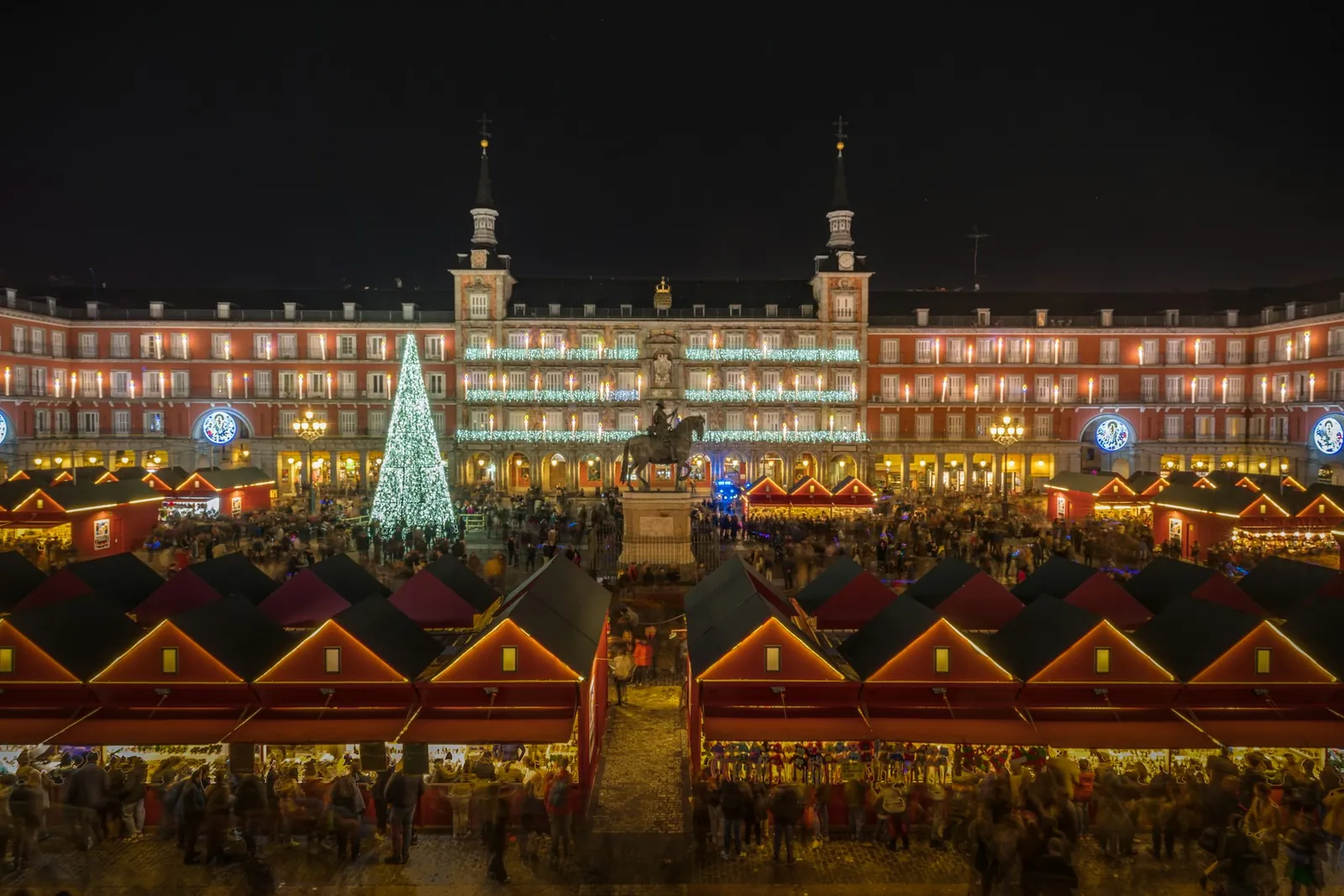 El mercado navideño de la Plaza Mayor de Madrid de noche, con un árbol iluminado y filas de puestos de techo rojo.
