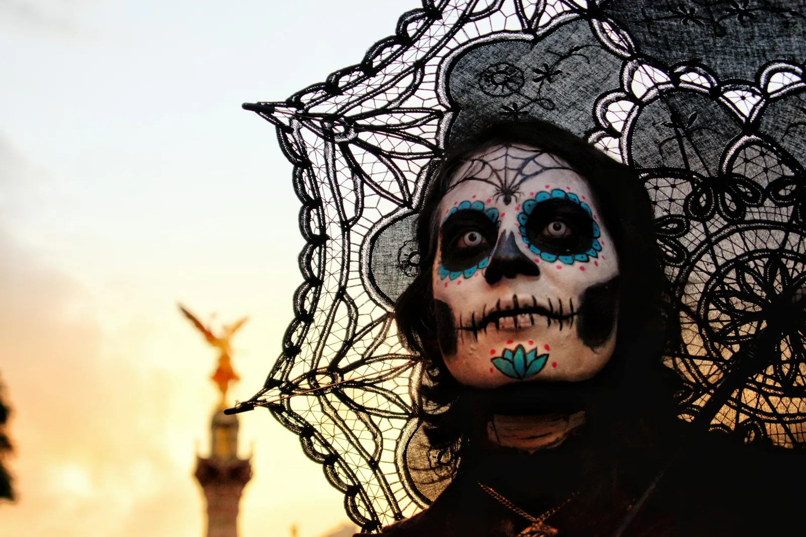 A woman in Catrina skull face paint, with dark eyes rimmed in turquoise, under a black lace parasol in Mexico City, the Angel of Independence behind her at sunset.