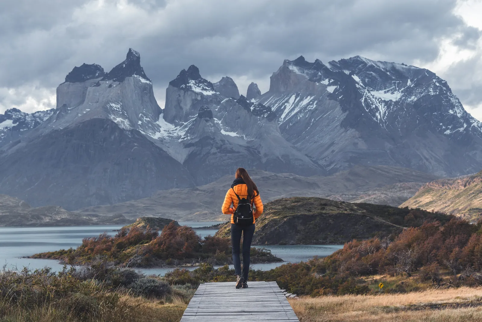 A hiker in an orange jacket walks a wooden boardwalk towards the granite peaks of Torres del Paine.