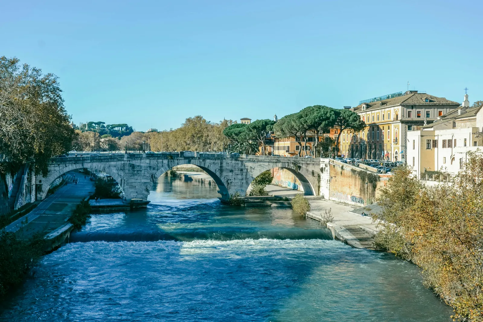 An old stone bridge crosses the Tiber to Tiber Island in Rome, with trees turning yellow along the river.