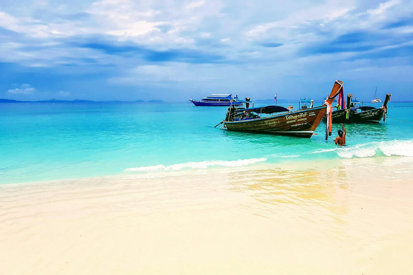 Dos barcas de cola larga de madera, con cintas de colores en la proa, flotan en agua turquesa y transparente frente a una playa blanca de las islas Phi Phi, con una persona nadando a su lado.