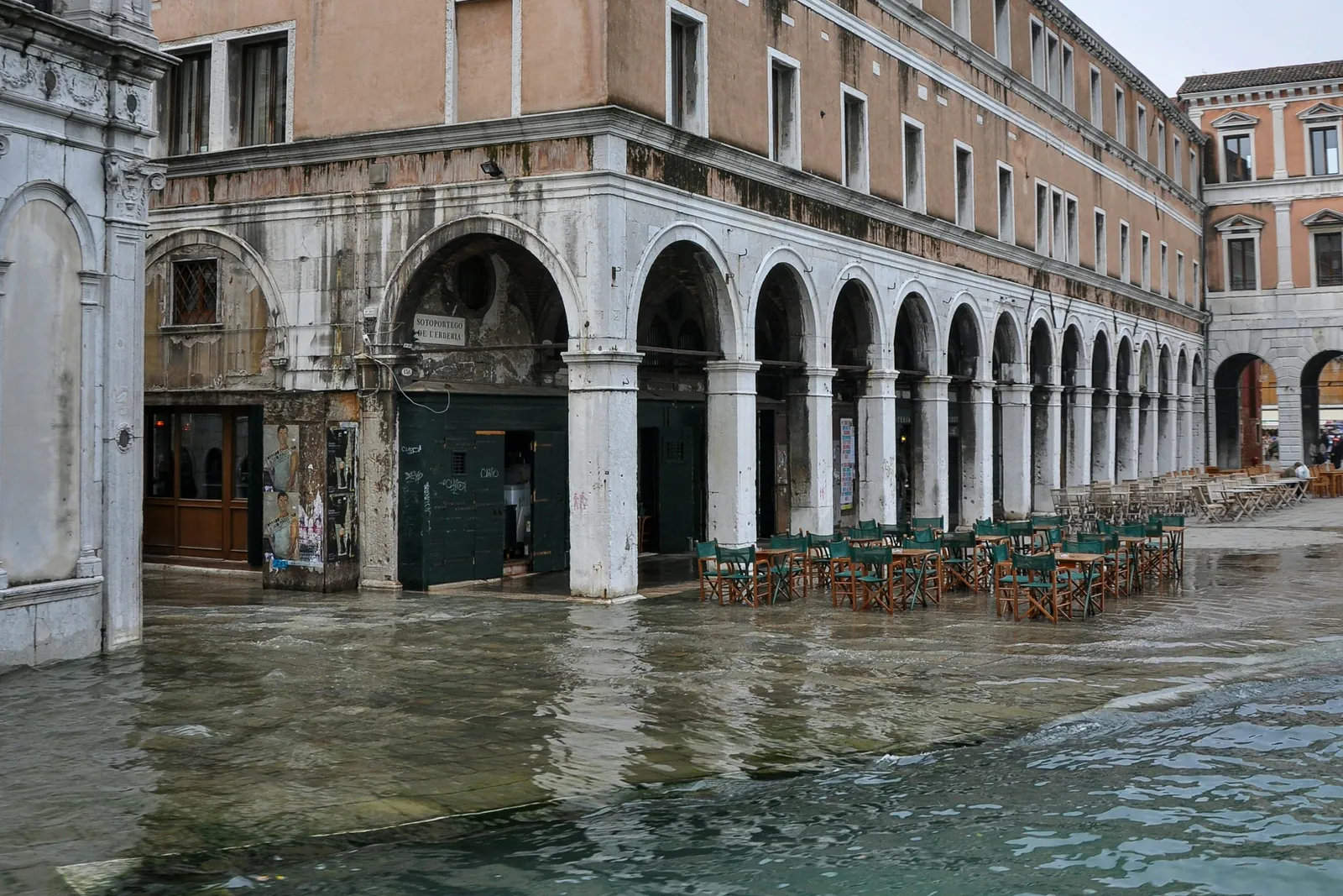 High water floods a square in Venice up to the arcades of an old building, with café tables standing in the water under a grey sky.