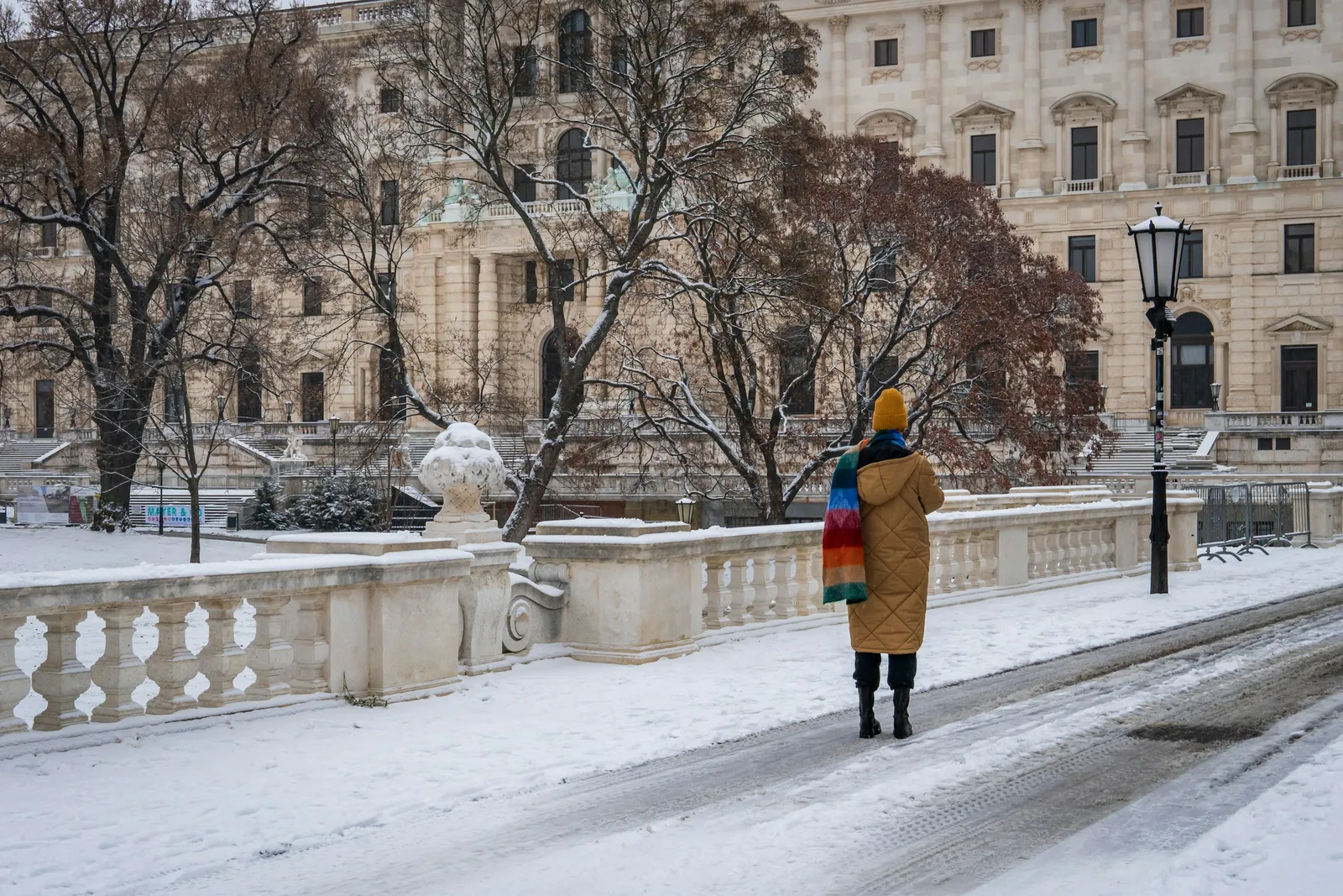 A person in a long winter coat and a striped scarf walks along a snowy street in Vienna, past a stone balustrade.