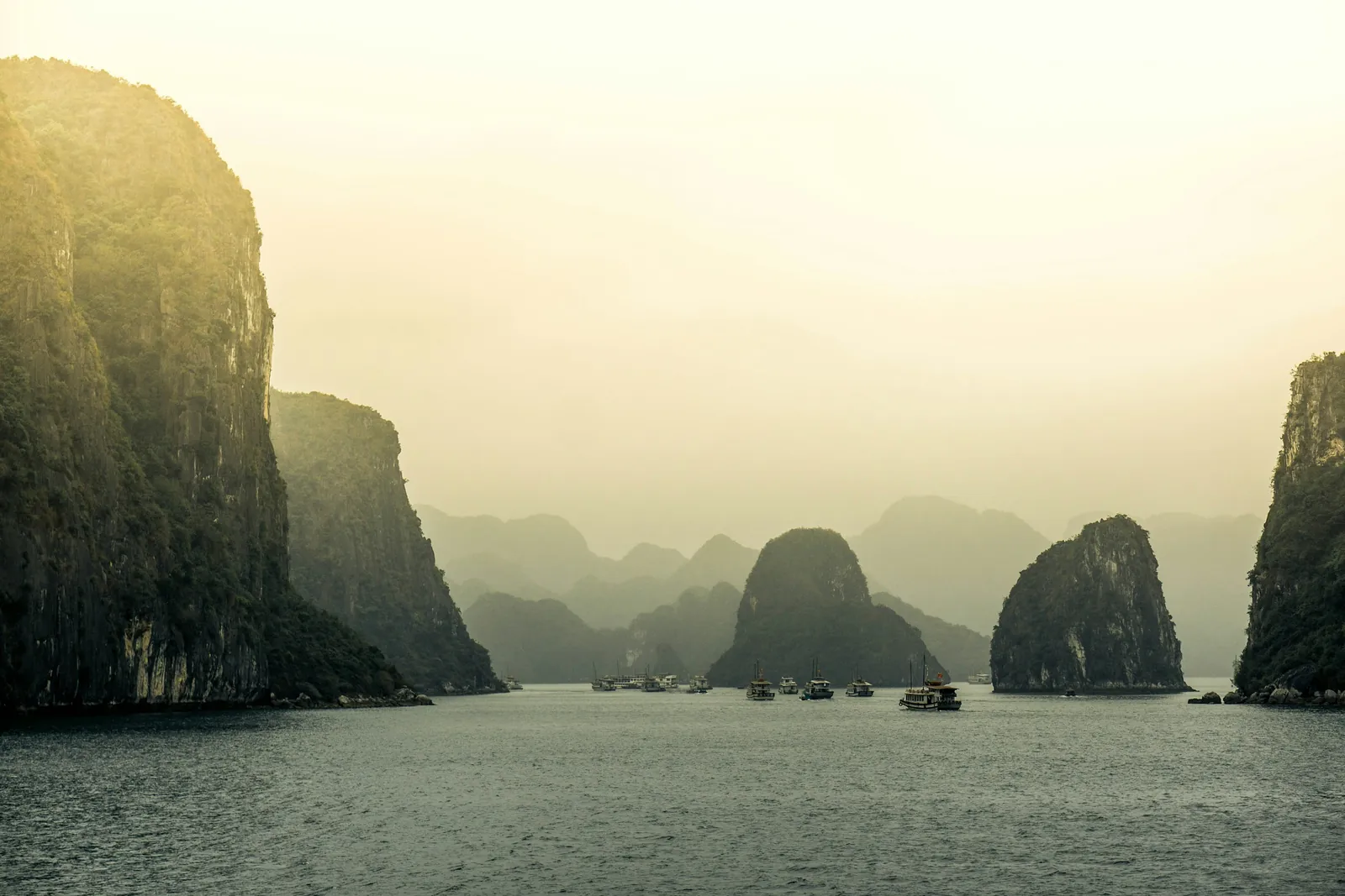 Limestone islands rise out of the mist over the calm water of Ha Long Bay, with small boats in the distance.
